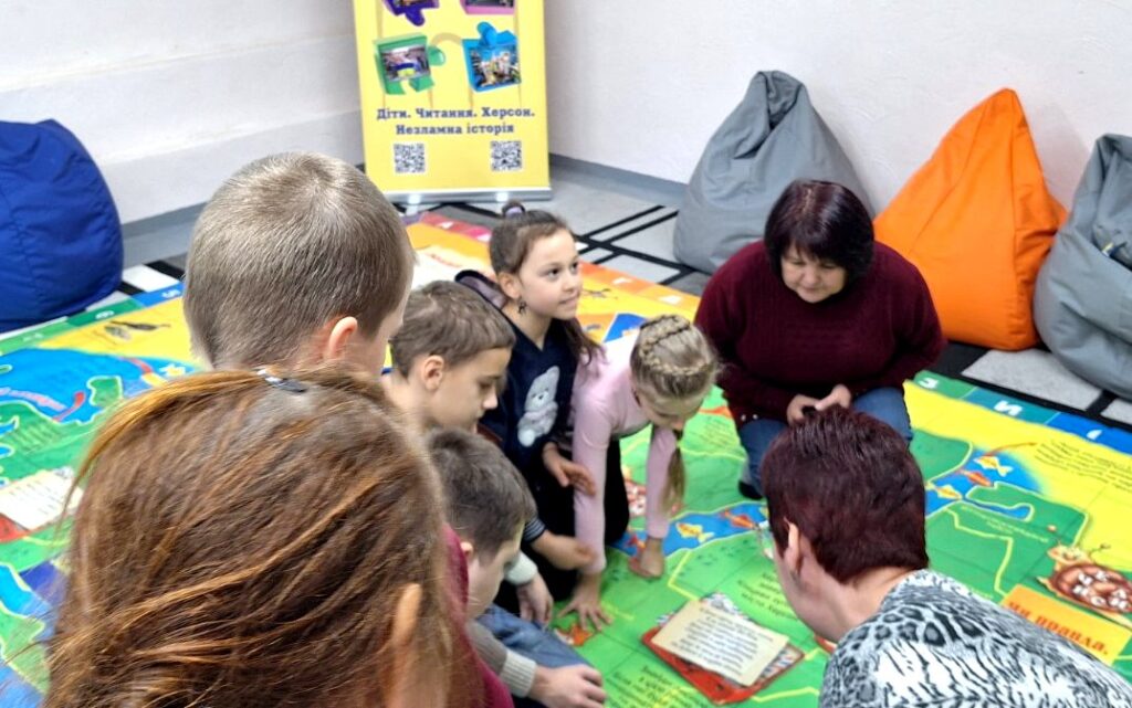 Діти й бібліотекарка грають на великому ігровому полі, розстеленому на підлозі