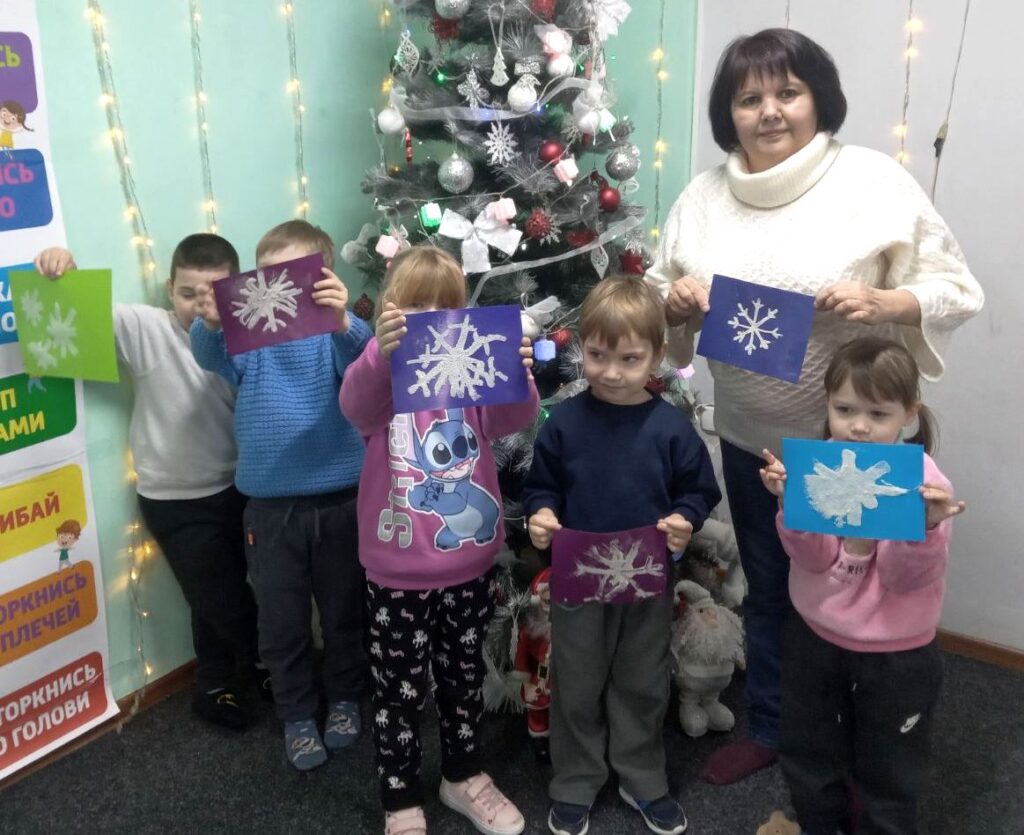 Учасники студії "Бібліомалята" демонструють свої малюнки сніжинок, стоячи перед прикрашеною різдвяною ялинкою