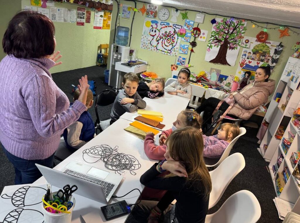 Діти в оздобленому малюнками приміщенні слухають розповідь бібліотекарки, сидячи довкола довгого столу