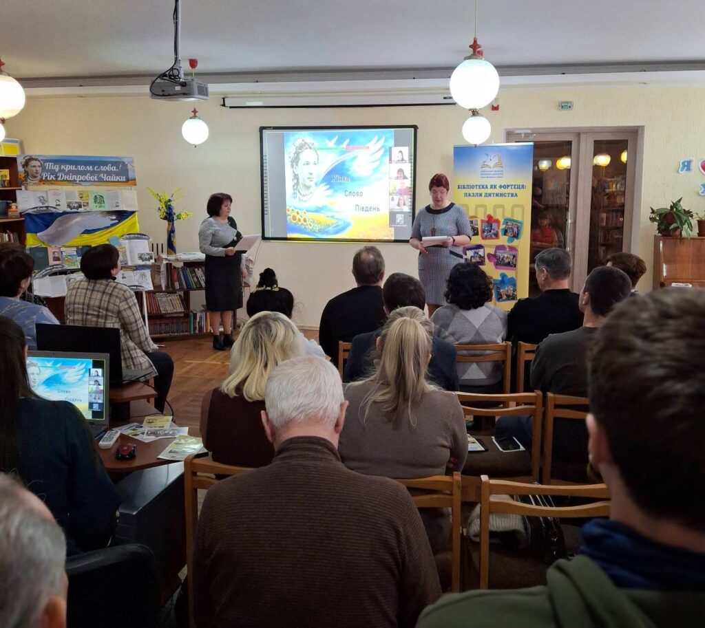 Учасники заходу у залі бібліотеки, біля великого екрану - дві ведучі