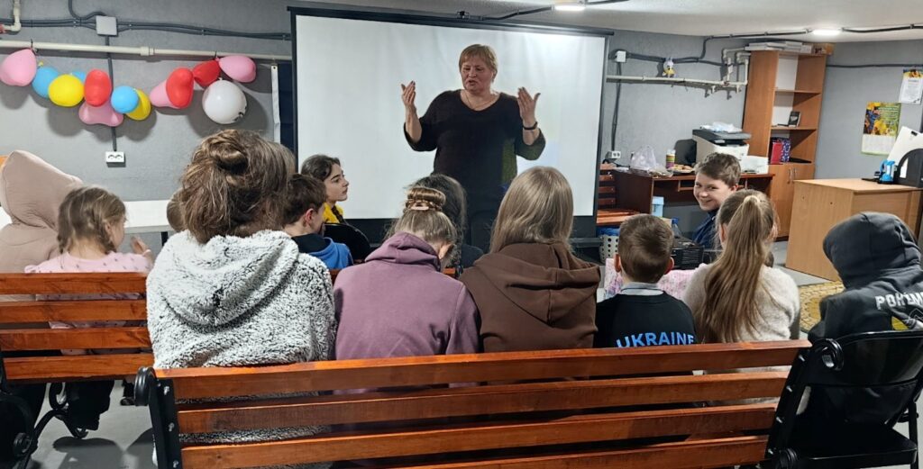 Учасники заходу слухають бібліотекарку, сидячи на довгих лавках в укритті