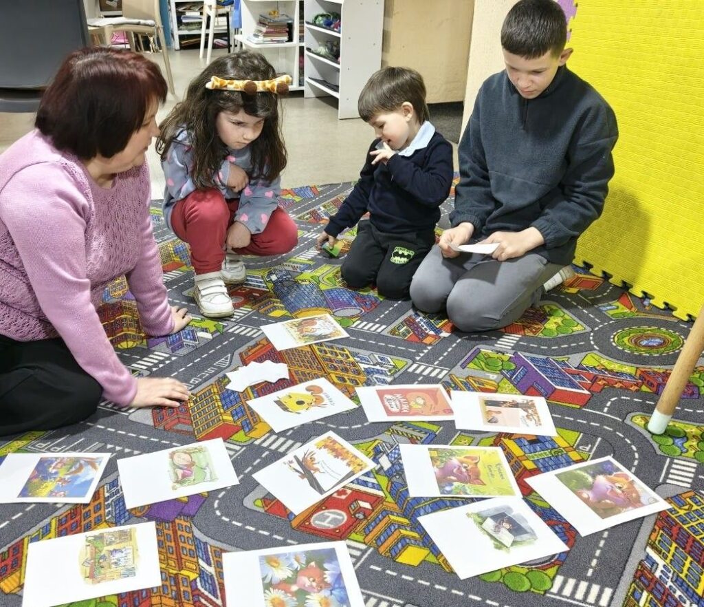 Учасники програми розглядають кольорові ілюстрації, сидячи на килимі поруч з бібліотекаркою