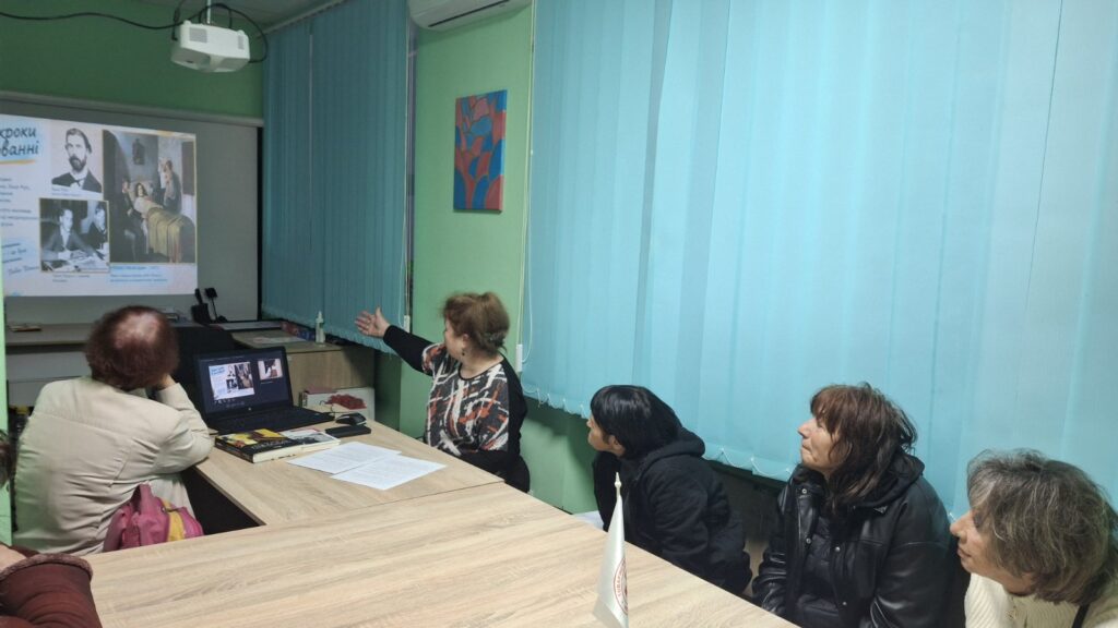 Учасники зустрічі слухають розповідь бібліотекарки, розглядаючи презентацію на великому екрані