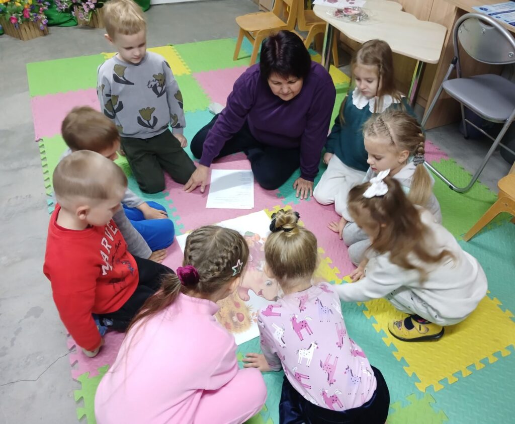 Діти сидять колом на килимку разом з бібліотекаркою, розглядаючи малюнки, - фото